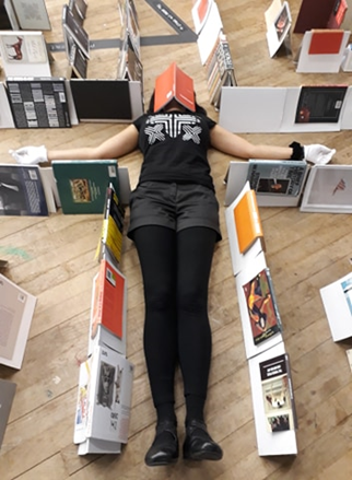A person lies on the ground with their arms outstretched and a book open on top of their face. Surrounding them are rows of books lying spine-up over A-shaped frames.