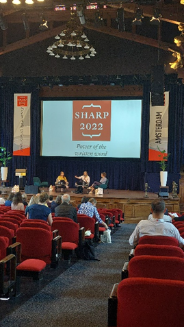 A panel presentation at the SHARP 2022 conference. There are three presenters on the stage, and the audience is pictured from behind.