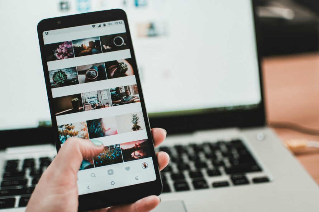 Person holding a smartphone in front of a laptop. Both are on and have pictures on screen. Photo by Lisa from Pexels.