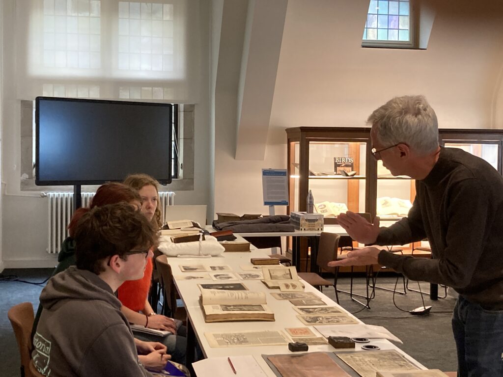 A curator stands in front of a table of rare books talking to a group of seated students.