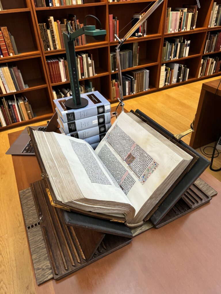 A green document camera, sitting on a stack of books, focuses on a Gutenberg Bible in a wooden book support placed beneath the camera on a table.