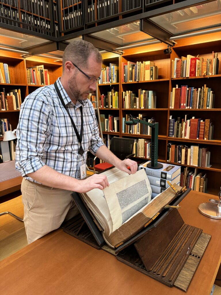 A white man in a blue and white shirt and khaki pants turns the page of a Gutenberg Bible. The book is in a wooden book support with a green document camera focusing on it from atop a stack of books.