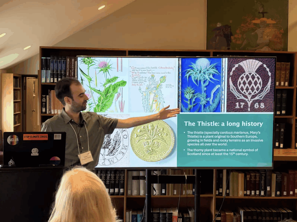 Photo of Pedro Toniazzo Terrez standing in front of a projected slideshow. He is pointing to a slide that shows "The Thistle: a long history" and contains four illustrations of thistles.