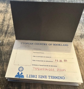 A Photograph of the first page of Bookland Passport with space for the holder’s photo, name, and date of birth. All are left empty, except for “Date of emission”, where an added stamp indicates “26 JUL 2024” and “Embassy of issue” which specifies “Typewronger Books” in handwriting. The page is titled “Utopian Country of Bookland” and reproduces the logo with the open book and terrestrial sphere as well as the slogan “Libri Sine Termino” at the bottom.