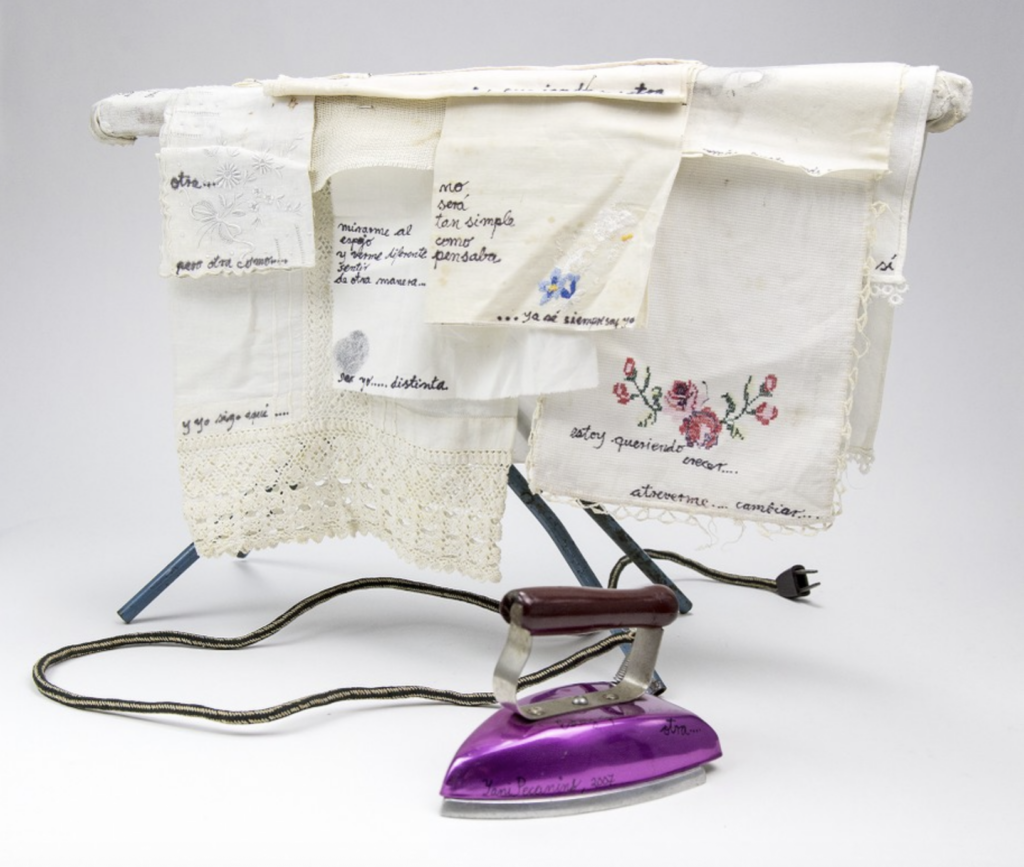 Artists’ book composed of child’s play ironing board and iron. The bright purple iron is on the foreground. The ironing board is pinned with light colored pieces of cloth, some with decorative edges, some that are lightly stained, that are further adorned with florals. Pieces bear written fragments in Spanish