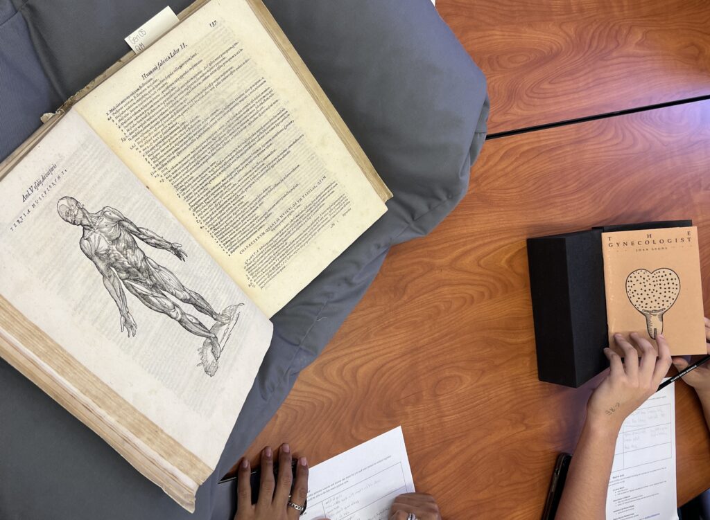 Overhead shot of two undergraduate student hands as they fill out a worksheet. In front of the dark-skinned student on the left is a copy of ‘Anatomia’ open to a diagram of the human muscles. The light-skinned student on the right’s has a hand on the cover of ‘The Gynecologist.' 