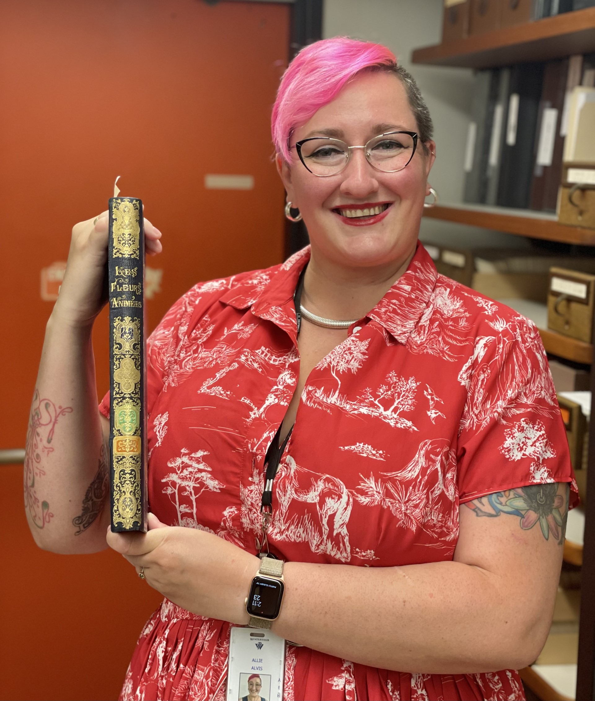 Allie, who has pink short hair and is wearing a red dress, holds an old book.