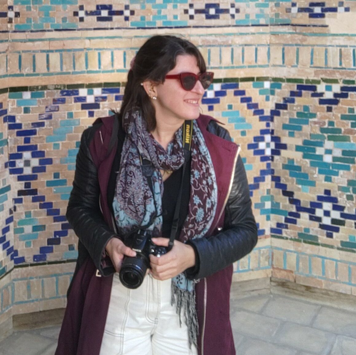 Julia is standing in front of a blue and beige tiled wall wearing a scarf, sunglasses, white jeans, and is holding a camera.