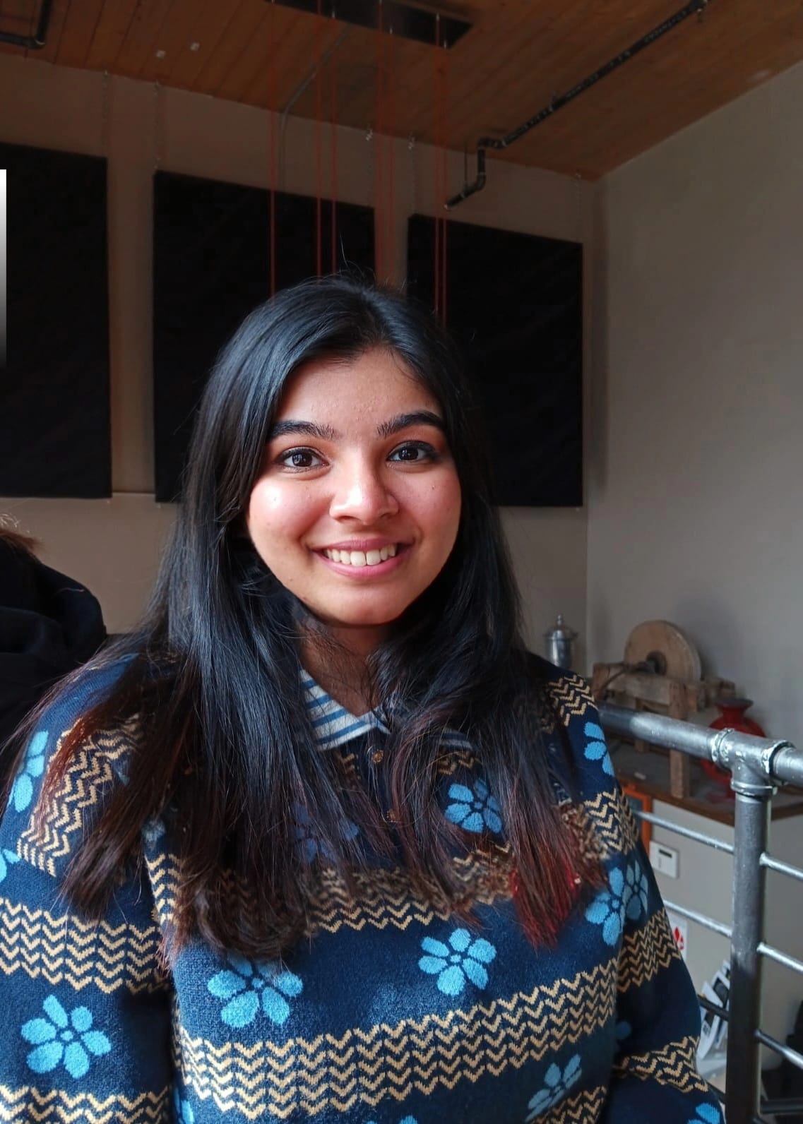 Anushmita is sitting in front of a white wall with three paintings on it wearing a colourful shirt.